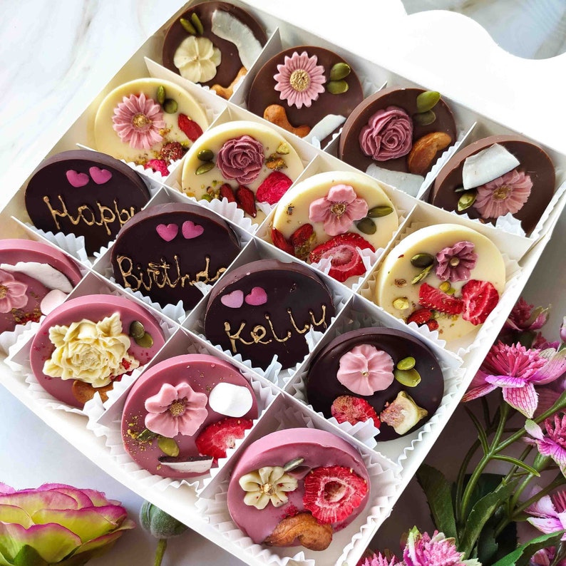 May include: A white box filled with assorted chocolate treats. The chocolates are decorated with various designs, including flowers, hearts, and the words "Happy Birthday" and "Kelly".
