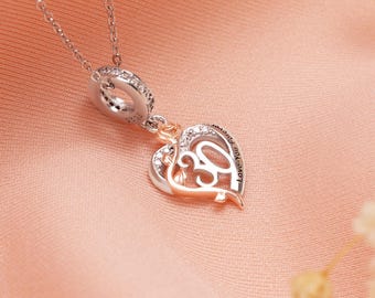 Puede incluir: Un collar plateado con un colgante en forma de corazón. El colgante presenta una rosa de oro rosa y el número 30, con las palabras "Love You Forever" inscritas en el lateral. El colgante está adornado con pequeños cristales transparentes.