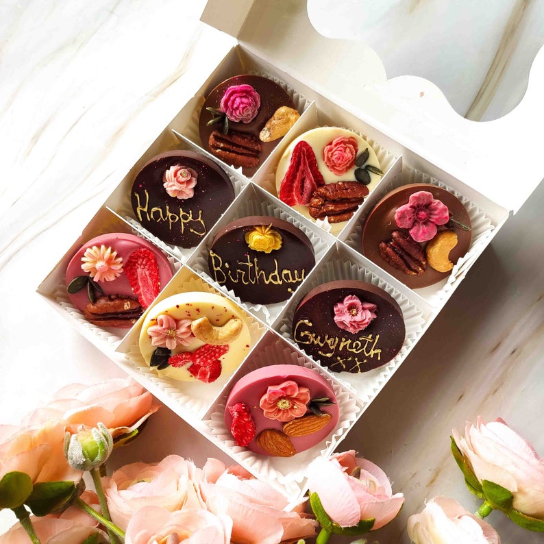 May include: A white box filled with nine chocolate-covered treats. Each treat is decorated with a different design and inscription. The inscriptions include "Happy Birthday", "Gwyneth", and "Happy".