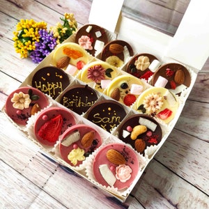 May include: A white box filled with assorted chocolate treats. The chocolates are decorated with various designs, including flowers, fruits, and words like "Happy" and "Birthday".
