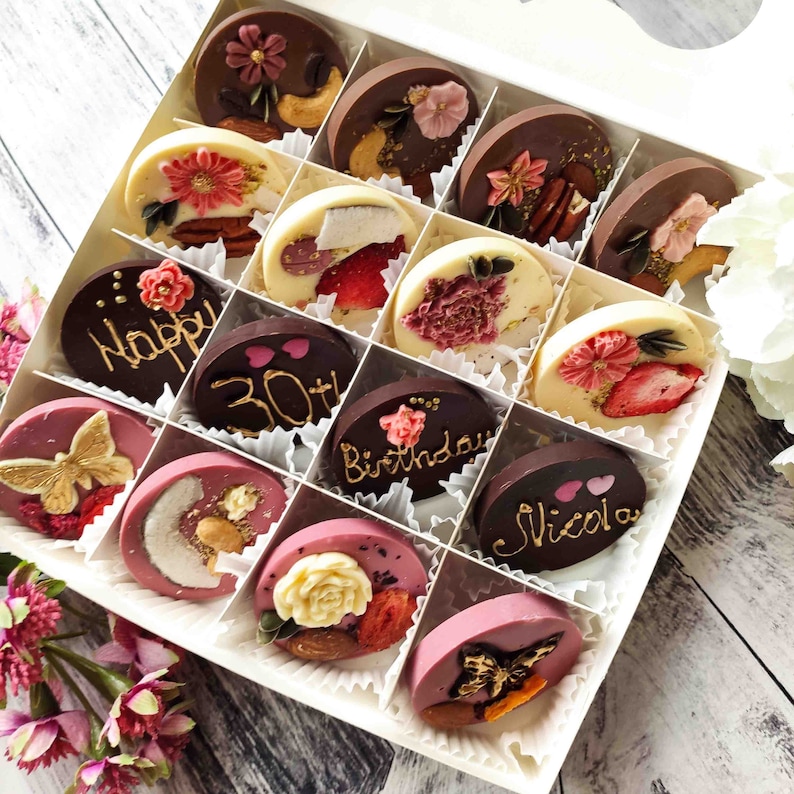 May include: A white box filled with 16 round chocolate discs. The chocolates are decorated with various designs, including flowers, butterflies, hearts, and text. The text on the chocolates includes "Happy 30th Birthday" and "Nicola".