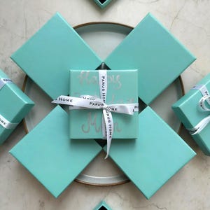 Puede incluir: Varias cajas de regalo turquesas, algunas con cintas blancas. La caja central est&aacute; envuelta con una cinta con la inscripci&oacute;n "PARUS HOME" y "Happy Mother's Day" en plata. Las cajas est&aacute;n dispuestas sobre una superficie neutra.