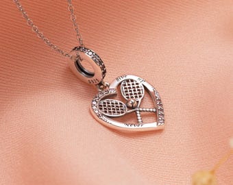 Puede incluir: Un collar con colgante plateado con temática de tenis. El colgante en forma de corazón presenta dos raquetas de tenis y las palabras "Give up Never". El collar tiene una cadena delicada y un cierre decorativo.
