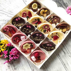 May include: A white box filled with 15 chocolate discs. The chocolates are decorated with various toppings, including nuts, dried fruit, and flowers. Some of the chocolates have messages written on them, such as "Happy 60th Birthday Donna".