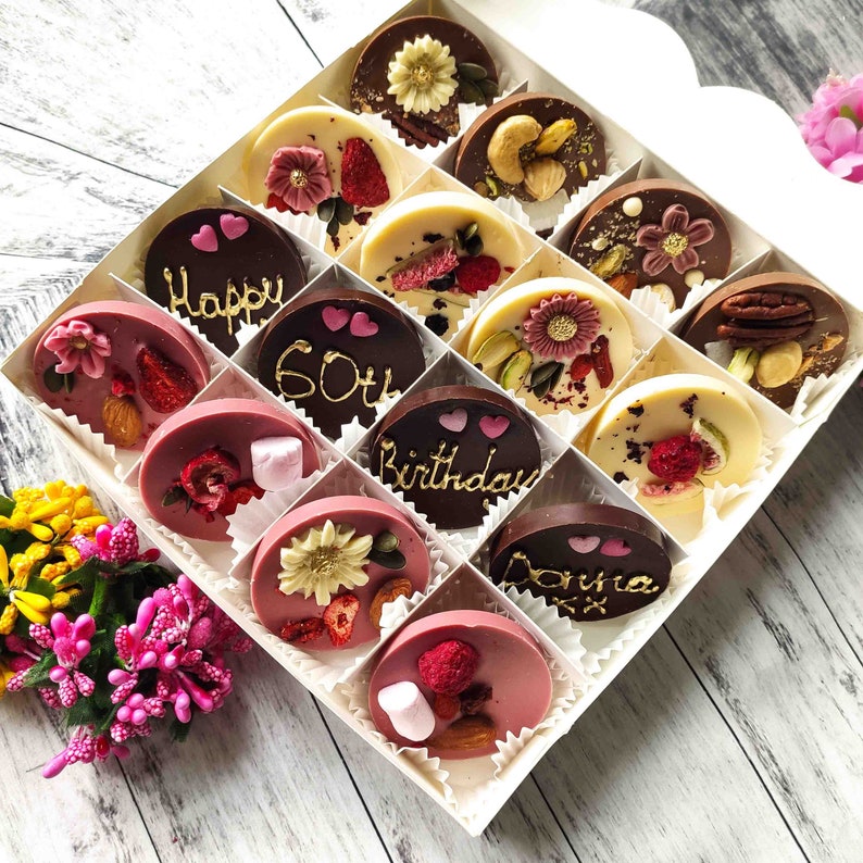 May include: A white box filled with 15 chocolate discs. The chocolates are decorated with various toppings, including nuts, dried fruit, and flowers. Some of the chocolates have messages written on them, such as "Happy 60th Birthday Donna".