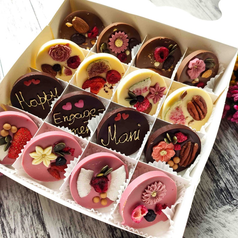 May include: A white box filled with 16 assorted chocolate discs. The chocolates are decorated with various toppings, including dried fruit, nuts, and sprinkles. Some of the chocolates have messages written on them, such as "Happy" and "Engaged".