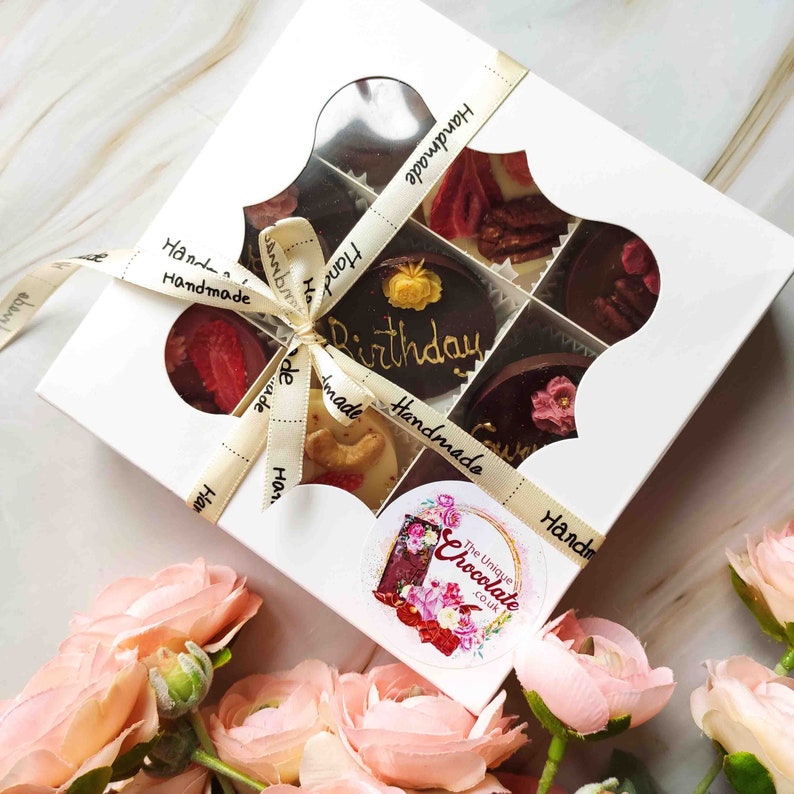 May include: A white box with a clear window showing a variety of handmade chocolates. The chocolates are arranged in a grid pattern and include a chocolate with the word "Birthday" written on it. The box is tied with a white ribbon and has the words "Handmade" printed on it.
