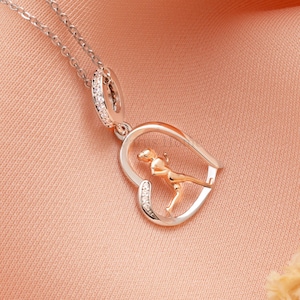 May include: A silver necklace with a heart-shaped pendant. The pendant features a gold-colored figure of a person in motion inside a silver frame. The frame is accented with small, clear stones. The necklace is displayed on a peach-colored surface.