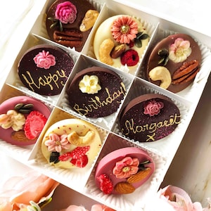 May include: Assortment of chocolate-covered treats in a white box. Each treat is decorated with a different design and message, including "Happy Birthday", "Happy", and "Margaret".