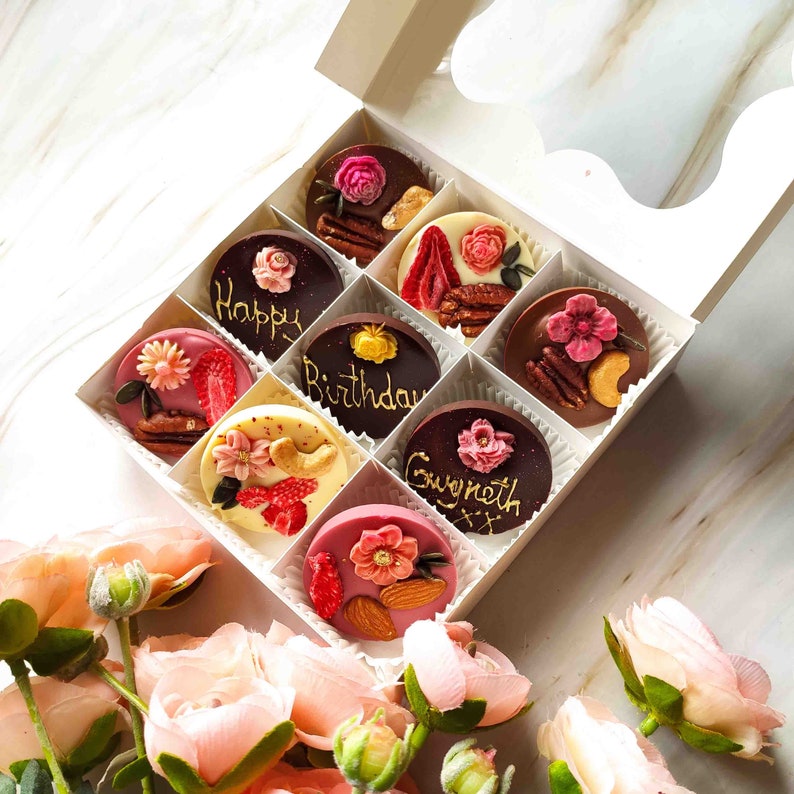 May include: A white box filled with eight chocolate-covered treats. Each treat is decorated with a different design, including flowers, nuts, and gold lettering. The lettering includes the words "Happy Birthday" and "Gwyneth".