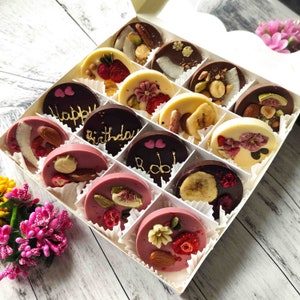May include: A white box filled with assorted chocolate discs. The chocolates are decorated with dried fruit, nuts, and gold lettering. Some of the chocolates have the words "Happy Birthday" and "Rob" written on them.