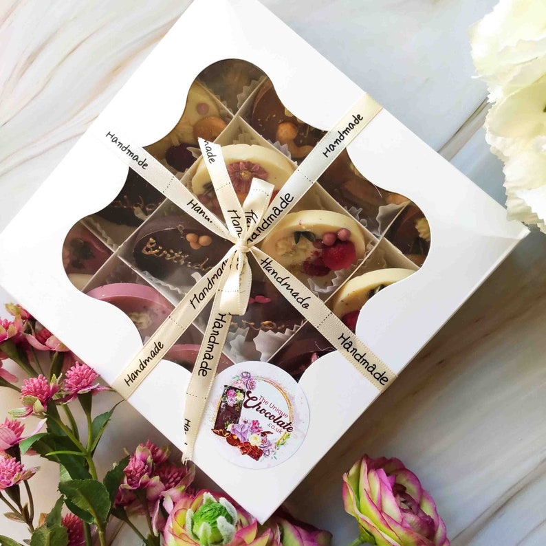 May include: A white box with a clear window showing a variety of handmade chocolates. The box is tied with a beige ribbon and has the words "Handmade" printed on each side. The box also has a sticker with the words "The Unique Chocolate Co. UK".