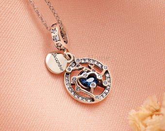 Puede incluir: Un collar plateado con un colgante con una piedra preciosa azul rodeada de cristales transparentes. El colgante tiene un diseño circular con la palabra "Diciembre" en un pequeño dije. El collar está sobre un fondo rosa.