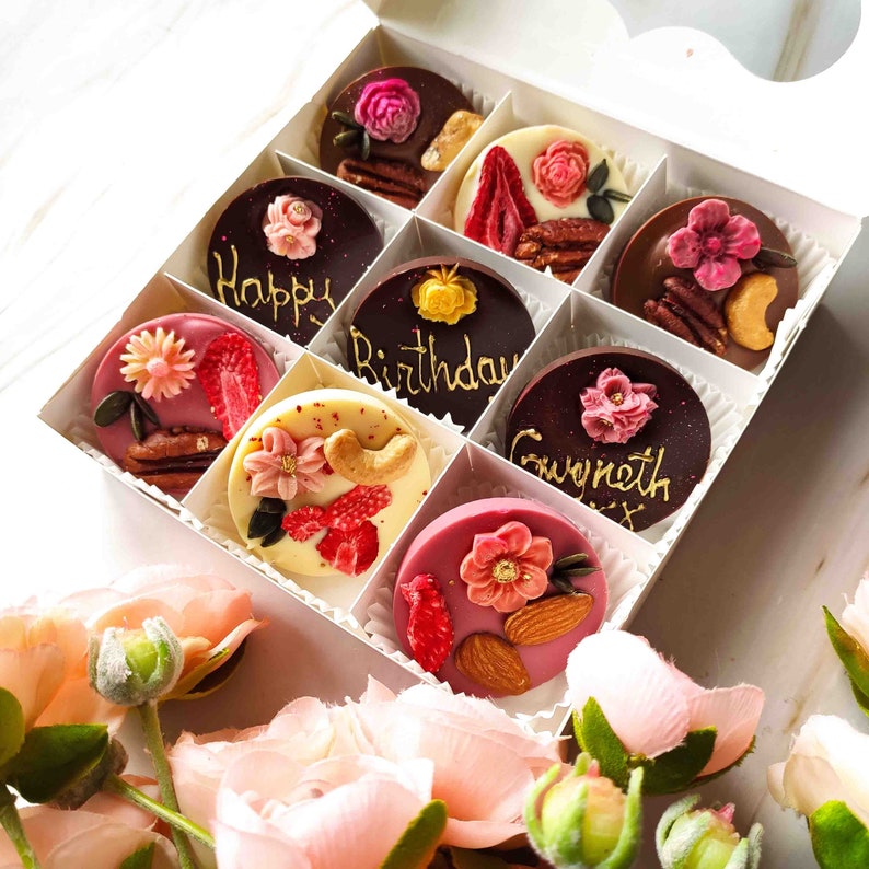 May include: A white box filled with nine chocolate-covered treats. Each treat is decorated with a different design, including flowers, nuts, and gold lettering. The lettering on the chocolates reads "Happy Birthday Gwyneth".