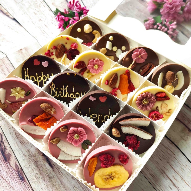 May include: A white box filled with 12 chocolate discs. Each disc is topped with different toppings, including dried fruit, nuts, and sprinkles. The box is decorated with the words "Happy Birthday" and "Friend".