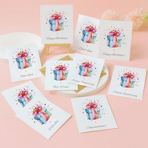 Puede incluir: Un conjunto de tarjetas de felicitaci&oacute;n blancas con ilustraciones de acuarela de cajas de regalo con lazos rosas. Las tarjetas tienen varios mensajes, incluyendo "Happy Birthday!", "Good Luck", "Best Wishes", "Happy Anniversary", "With Love" y "Congratulations!"