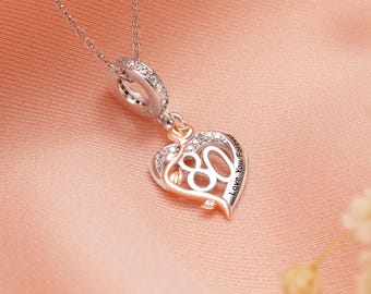 Puede incluir: Collar con colgante de corazón en plata y oro rosa con el número 80. El corazón está adornado con cristales transparentes y un detalle de rosa. La cadena del collar es plateada, sobre un fondo rosa suave.