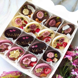 May include: A box of assorted chocolate treats with various designs and flavors. The chocolates are arranged in a grid pattern and feature decorative elements like flowers, hearts, and text. The text on the chocolates includes "Happy", "Birthday", and "Kelly".