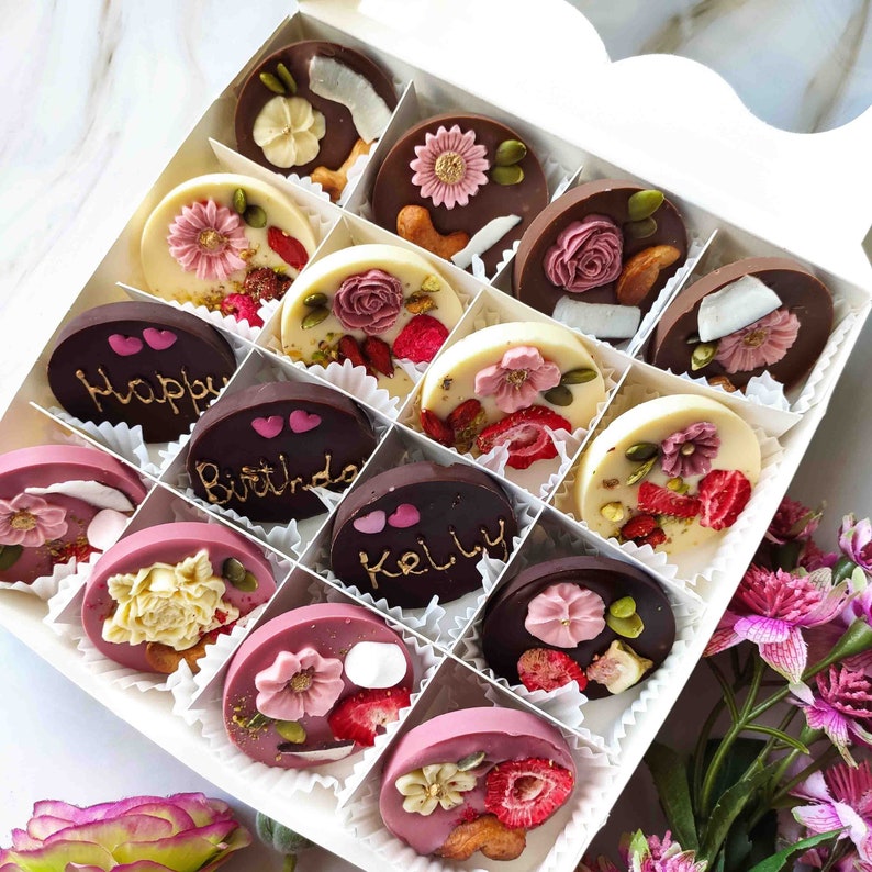 May include: A box of assorted chocolate treats with various designs and flavors. The chocolates are arranged in a grid pattern and feature decorative elements like flowers, hearts, and text. The text on the chocolates includes "Happy", "Birthday", and "Kelly".