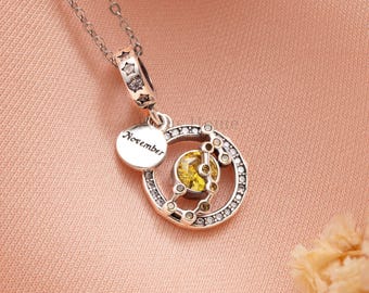 May include: A silver necklace with a pendant featuring a yellow gemstone surrounded by a circle of clear stones. A small disc with the word "November" is attached. The necklace has a delicate chain and a star-shaped clasp.