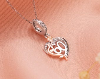 Puede incluir: Collar plateado con un colgante en forma de corazón. El colgante presenta el número "60" y las palabras "Love You Forever". El corazón está adornado con cristales transparentes y un diseño de rosa. El collar se muestra sobre tela rosa.