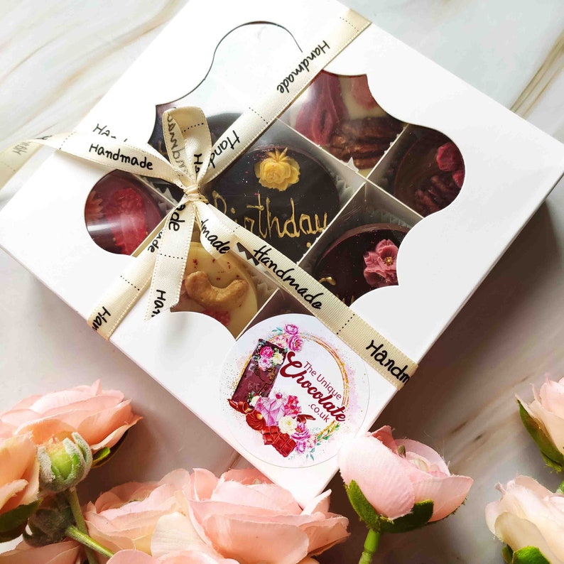 May include: A white box with a clear window, filled with six assorted chocolate treats. The box is tied with a gold ribbon with the words "Handmade" printed on it. The chocolates are decorated with flowers, sprinkles, and the words "Happy Birthday". The box has a sticker with the words "The Unique Chocolate.co.uk" on it.