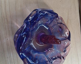 以下が含まれることがあります： ユニークな有機的な形状の手作りのガラスアート作品。青と紫の色合いが混ざり合い、まだらのキラキラとした効果があります。濃い赤色の円筒形の要素が中央から伸びています。ガラスは半透明で、その曲線に光が反射しています。