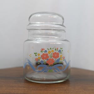 May include: A clear glass jar with a matching lid, featuring a colorful floral bouquet graphic in shades of coral, yellow, and blue, accented by a light blue ribbon design wrapped around the base of the jar, creating a decorative storage container.