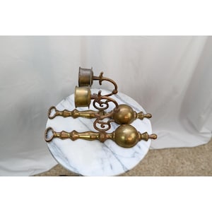 May include: Two matching brass candle holders featuring a long, slender stem with a rounded bulbous base and a ring handle at the end. Each holder has a curved, ornate arm supporting a cylindrical cup designed to hold a single taper candle.
