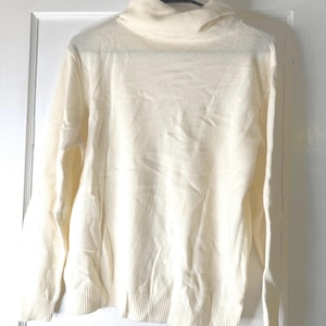 Könnte beinhalten: Ein cremefarbener Rollkragenpullover hängt an einem schwarzen Kleiderbügel vor einer weißen Tür. Der Pullover hat lange Ärmel und einen gerippten Saum und Bündchen. Der Rollkragen ist umgeschlagen. Der Stoff scheint ein weiches Strickmaterial zu sein.