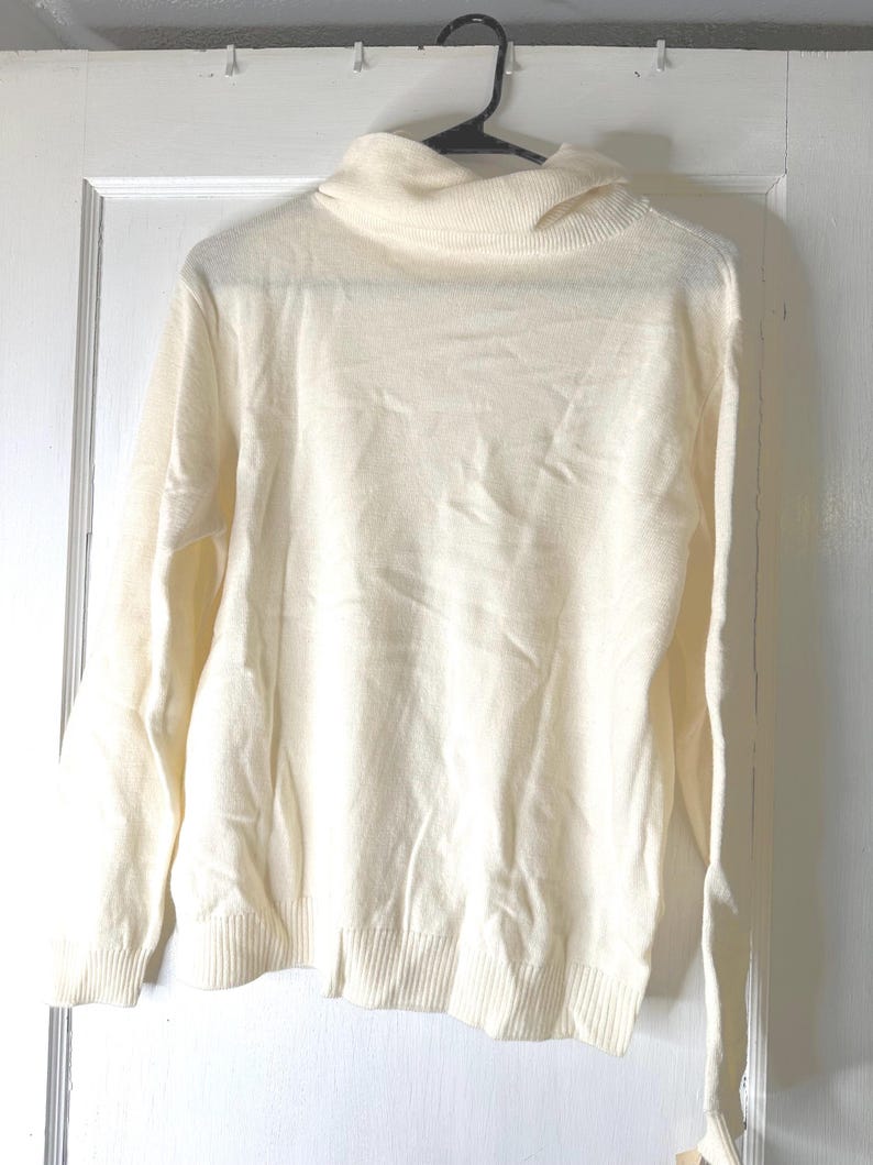 May include: A cream-coloured turtleneck sweater hangs on a black hanger against a white door. The sweater has long sleeves and a ribbed hem and cuffs. The turtleneck is folded over. The fabric appears to be a soft knit.