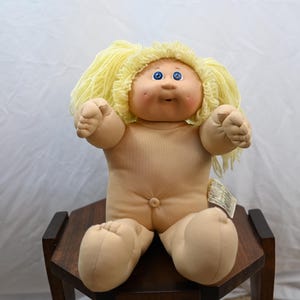 May include: A soft-sculpture doll with a round, peach-toned face, dimpled cheeks, and bright blue eyes. The doll features light yellow yarn hair styled in two pigtails and has a fabric body with a visible belly button. A small white tag is attached.