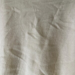 May include: Close-up of a textured, off-white fabric. The material has a subtle, woven pattern with visible folds and creases, creating shadows and variations in tone. The fabric appears soft and possibly lightweight.