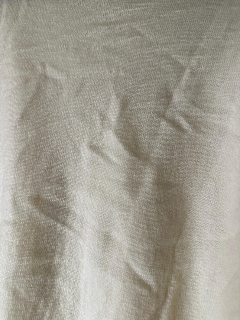 May include: Close-up of a textured, off-white fabric. The material has a subtle, woven pattern with visible folds and creases, creating shadows and variations in tone. The fabric appears soft and possibly lightweight.