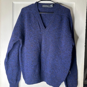 Könnte beinhalten: Ein blauer V-Ausschnitt-Pullover mit gesprenkeltem Muster, der an einem schwarzen Kleiderbügel hängt. Der Pullover hat gerippte Bündchen und einen gerippten Saum. Das Etikett lautet "Connemara Hams".