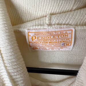 May include: Close-up of a cream-coloured Pendleton sweater. The sweater has a high collar and a tag with the Pendleton logo and text. The tag reads "Warranted to be a Pendleton" and "100% Virgin Wool". The sweater is a size M.