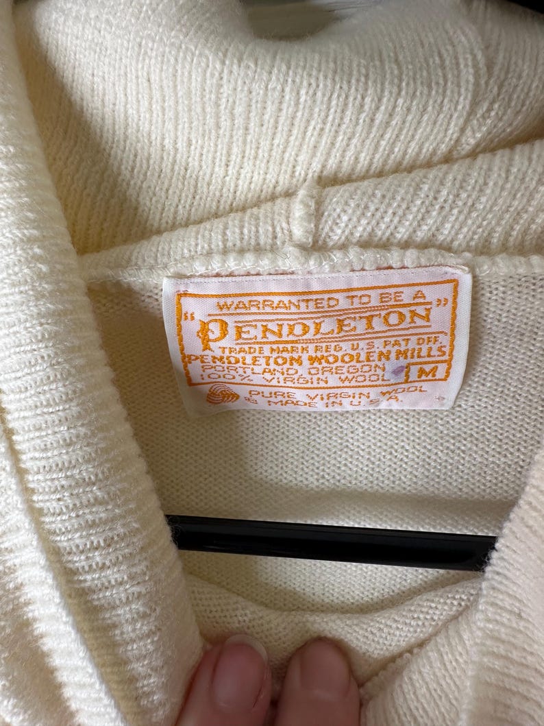 May include: Close-up of a cream-coloured Pendleton sweater. The sweater has a high collar and a tag with the Pendleton logo and text. The tag reads "Warranted to be a Pendleton" and "100% Virgin Wool". The sweater is a size M.