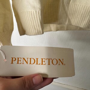 May include: Cream-coloured sweater with ribbed cuffs and hem. A tag with the brand name "PENDLETON" in orange lettering is held in front of the sweater.
