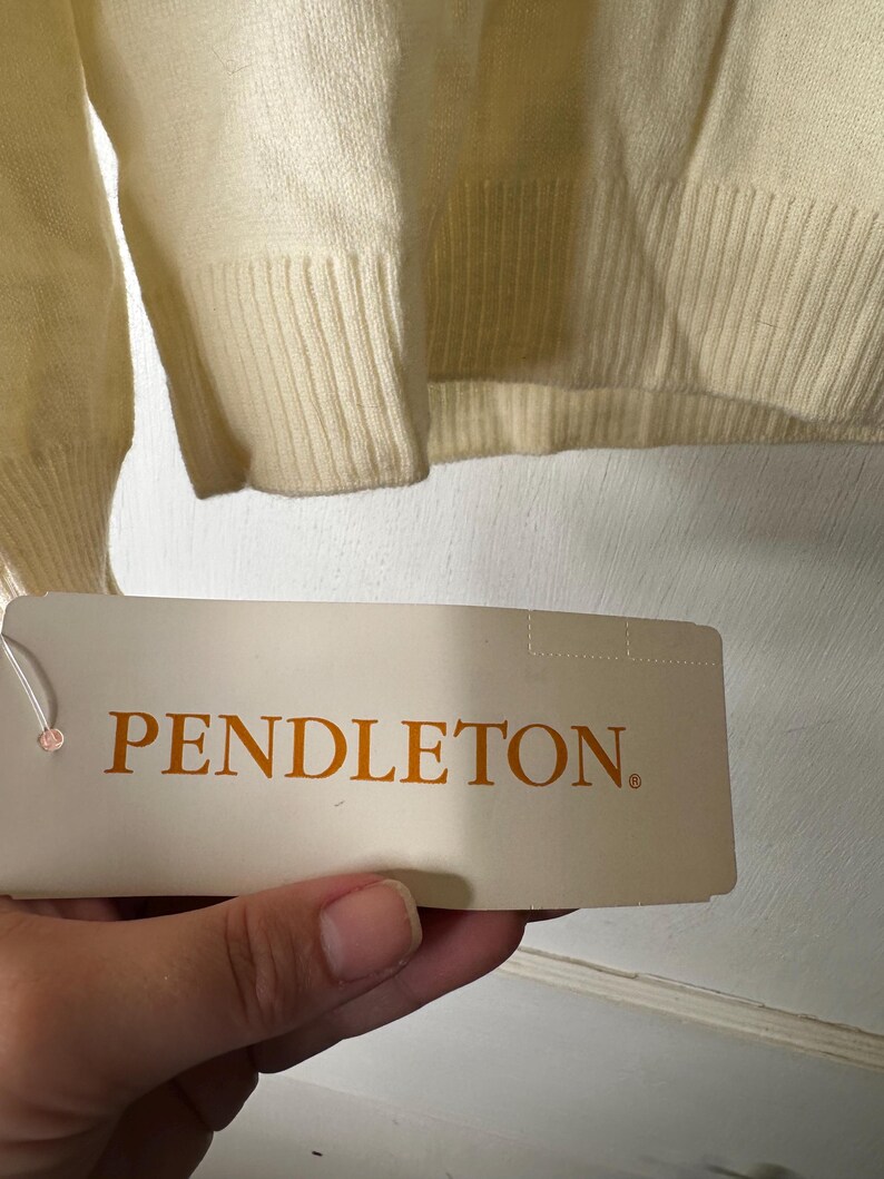 May include: Cream-coloured sweater with ribbed cuffs and hem. A tag with the brand name "PENDLETON" in orange lettering is held in front of the sweater.