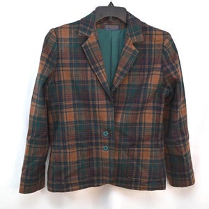 Puede incluir: Una chaqueta de manga larga con un patrón de cuadros en marrón, verde oscuro y azul marino. La prenda tiene solapas de muesca, un forro interior de color verde oscuro sólido y dos botones a juego en el cierre frontal. Se muestra en una percha negra.