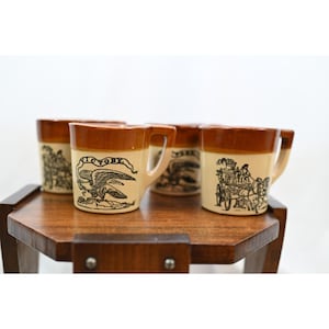Puede incluir: Un juego de cuatro tazas de cerámica con un diseño bicolor que presenta una base de color crema y un borde marrón. Cada taza muestra una ilustración distinta en tinta negra, incluyendo un águila con el texto VICTORY y una carroza.
