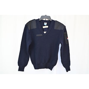 May include: A dark navy blue V-neck sweater featuring black fabric shoulder patches and a black rectangular patch on the left sleeve. The sleeve patch displays a small horizontal tricolor flag emblem in red, white, and blue. It has long sleeves.
