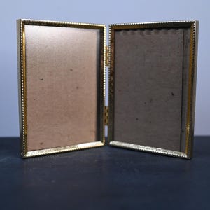 May include: A gold-colored, hinged picture frame, open to display two brown backing boards. The frame has a beaded edge design. The frame is on a dark surface with a light blue background.
