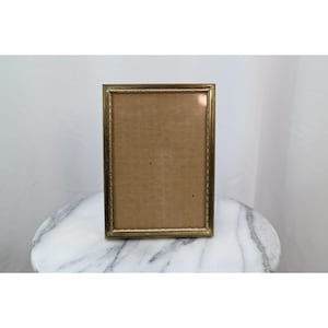 May include: A rectangular picture frame with a metallic gold finish and a textured, beaded border design. The frame features a clear glass front and a plain, light brown backing insert, presented in a vertical orientation for display.
