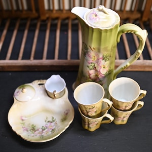 Puede incluir: Un juego de té vintage con una jarra alta verde con tapa, una bandeja para servir con una pequeña crema y cuatro tazas de té. El juego está decorado con motivos florales rosas y detalles dorados, sugiriendo un estilo delicado y antiguo.