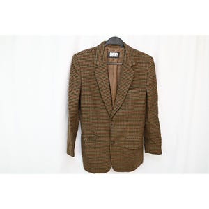 May include: A brown and green houndstooth blazer featuring a notched lapel, two front buttons, and two flap pockets. The interior is lined in brown fabric with a black DKNY brand label at the neck. The blazer has long sleeves and a structured fit.
