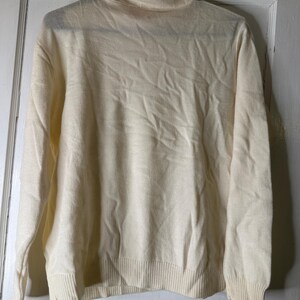 May include: Cream-coloured turtleneck sweater with long sleeves. The sweater has a ribbed hem and cuffs. The garment is hanging on a hanger against a white door. A tag is attached to the sleeve.