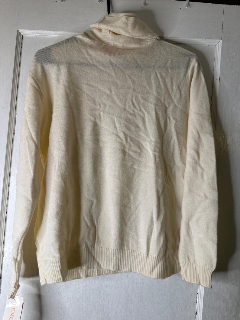 May include: Cream-coloured turtleneck sweater with long sleeves. The sweater has a ribbed hem and cuffs. The garment is hanging on a hanger against a white door. A tag is attached to the sleeve.