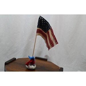 May include: A small fabric American flag attached to a thin wooden dowel. The flag features a blue canton with white stars and alternating red and off-white stripes. The dowel is secured in a small, speckled white base with red and blue fabric.