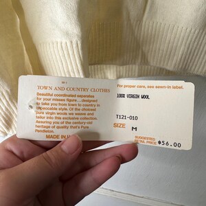 May include: Cream-coloured sweater with a ribbed hem and cuffs. A tag reads "TOWN AND COUNTRY CLOTHES" and "100% VIRGIN WOOL". The tag also indicates size M and a suggested retail price.